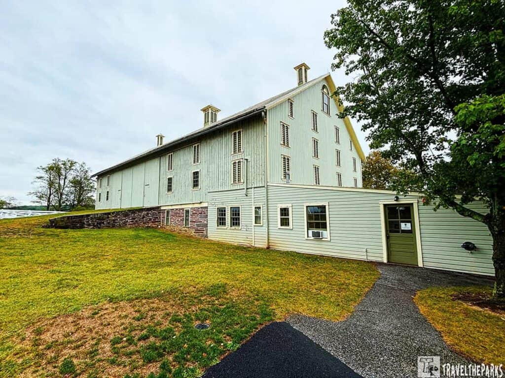 a large, light green barn with vertical siding, located at the Eisenhower National Historic Site. It features an attached structure that was once a milk house, now converted into a Secret Service office. The barn has a pitched roof with two cupolas, and numerous tall, narrow windows. The foundation is made of stone, contrasting with the wooden upper structure. In the foreground, there is a path leading to a doorway in the attached building, and the area around the barn is a well-maintained grassy lawn. A tree with dense foliage is visible on the right side, and the sky is overcast.