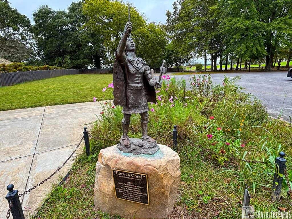 Bronze statue of "Etowah Chief" on a stone pedestal, surrounded by flowers and grass.

