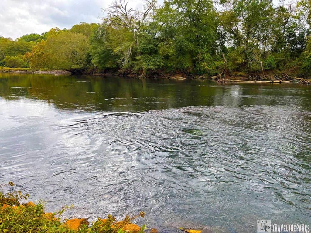 Etowah River with gentle ripples near a rock wier and wooded banks under a cloudy sky.

