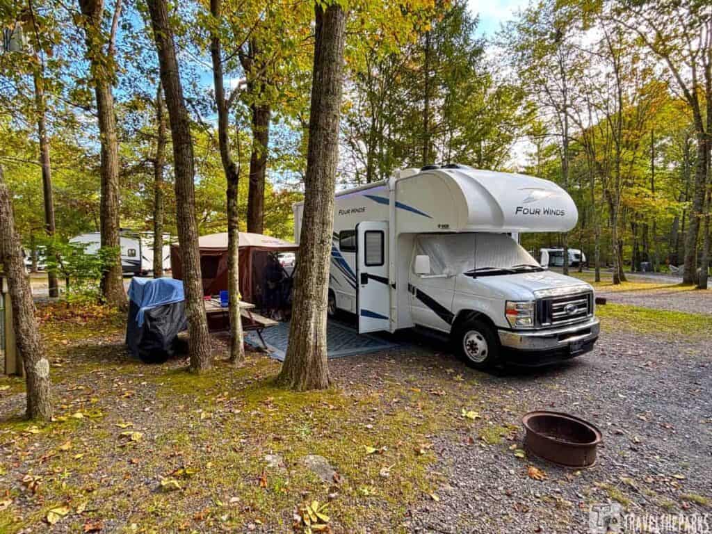 Campsite with a "Four Winds" RV, a canopy tent, and a metal fire pit at a wooded campground.

