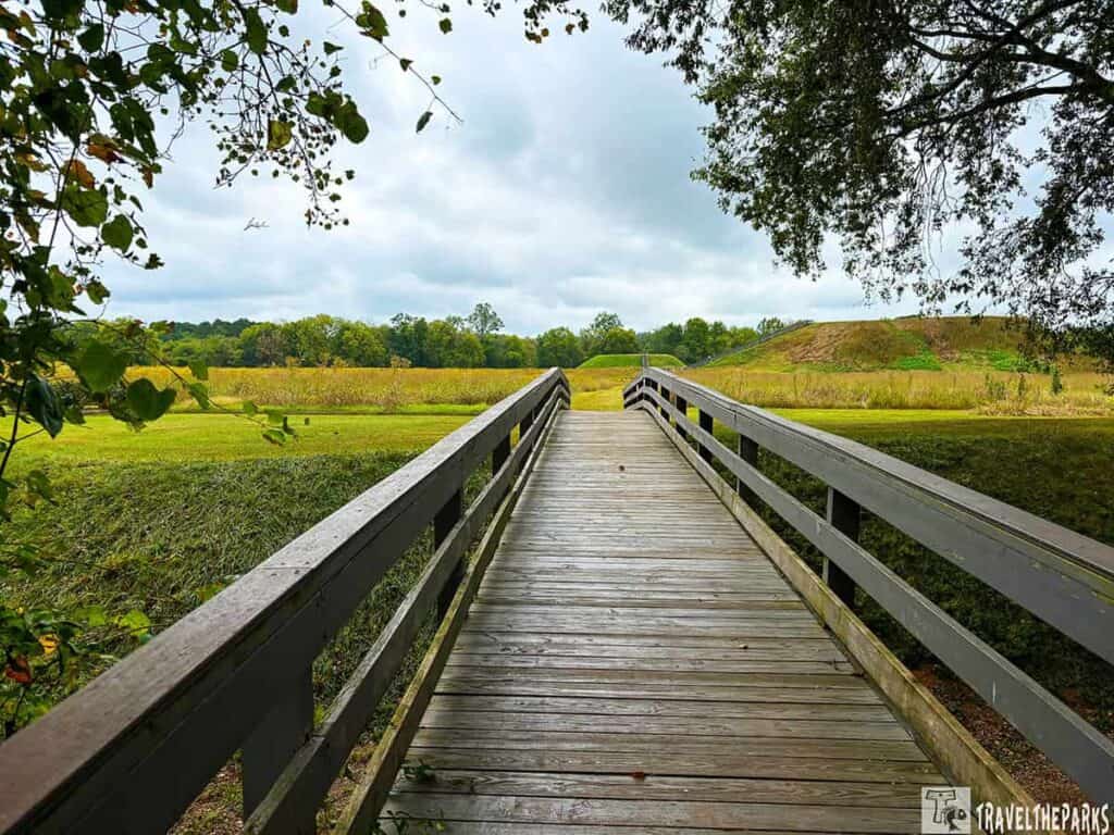 A wooden boardwalk leads through grassy fields towards the Etowah Mounds under an overcast sky.

