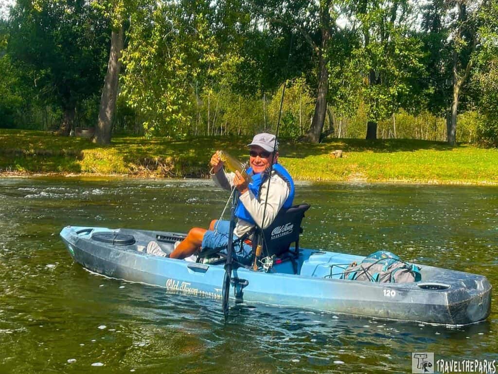 A person in a blue kayak holds up a fish on a river, with trees in the background.
