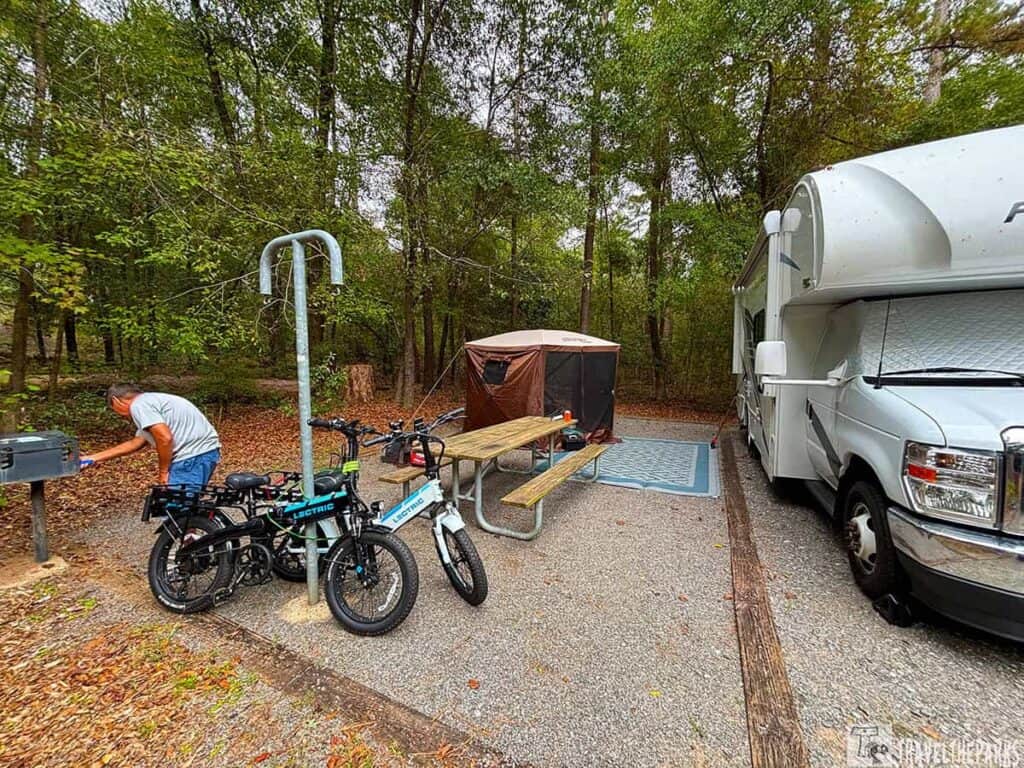 Campsite with RV, picnic table, tent, electric bikes, and a man grilling. Surrounded by forest.

