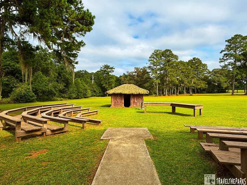 A small wooden hut with a thatched roof sits in a grassy field at Kolomoki Mounds State Park, surrounded by wooden benches and picnic tables, with trees in the background.