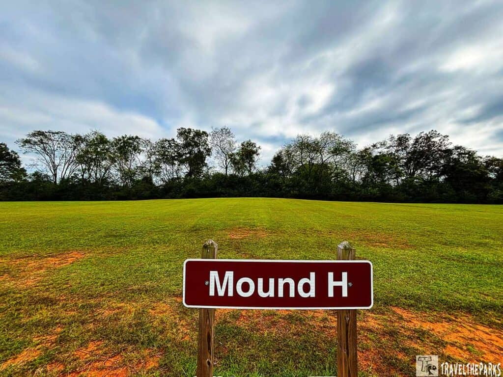 A sign labeled "Mound H" in front of a grassy field with trees and a cloudy sky.

