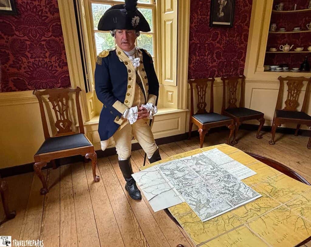 A person in 18th-century military attire stands in a historic room by a table with a large map.