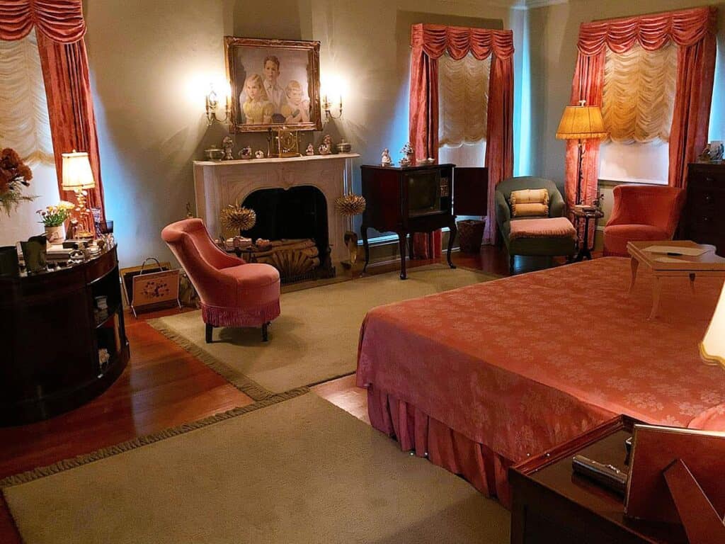 A traditionally decorated master bedroom with a pink bedspread, white fireplace, and brass sconces. Two windows feature pink wavy curtains, and various upholstered chairs furnish the space. Eisenhower Homestead.