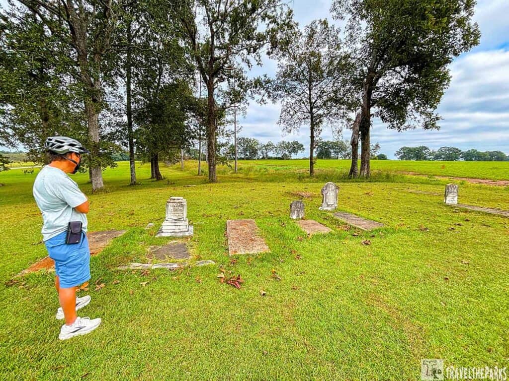 Person in helmet observing gravestones in a grassy area with trees and open field in the background.