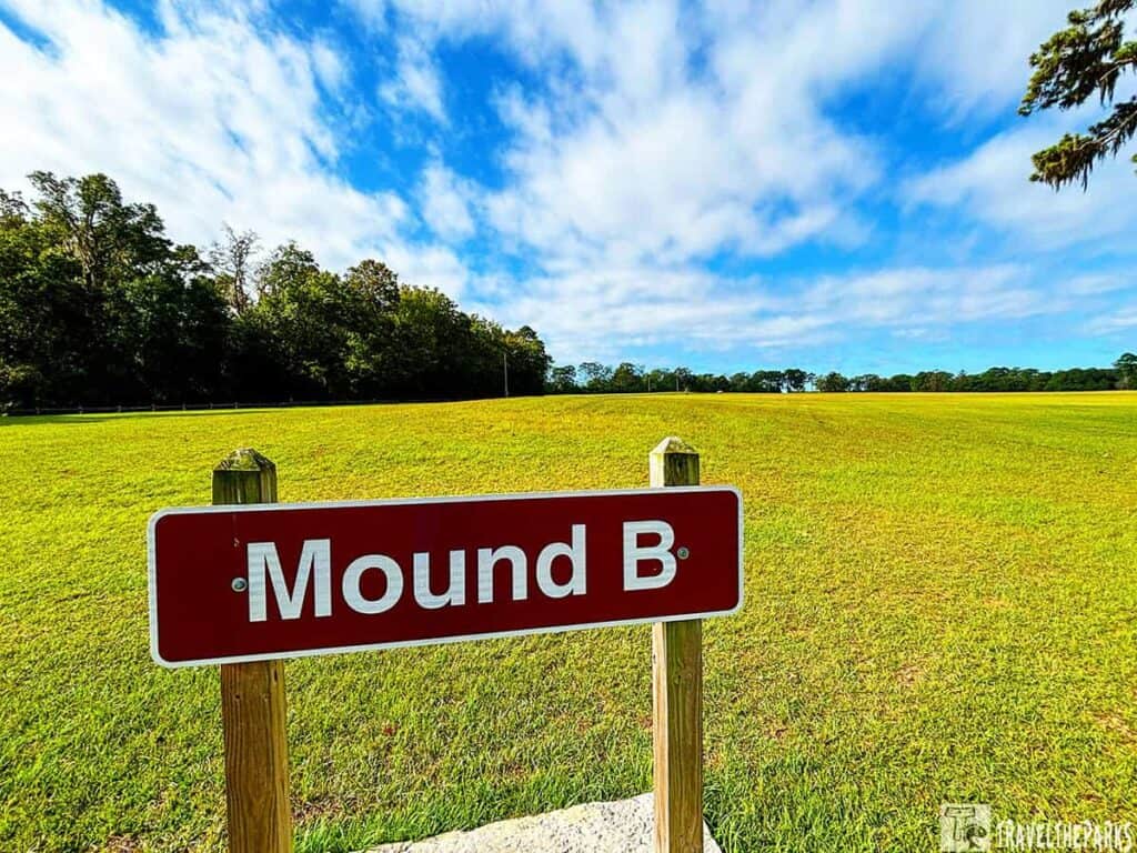 A red sign reading "Mound B" at a grassy field in Kolomoki Mounds State Park with a tree line and cloudy sky.


