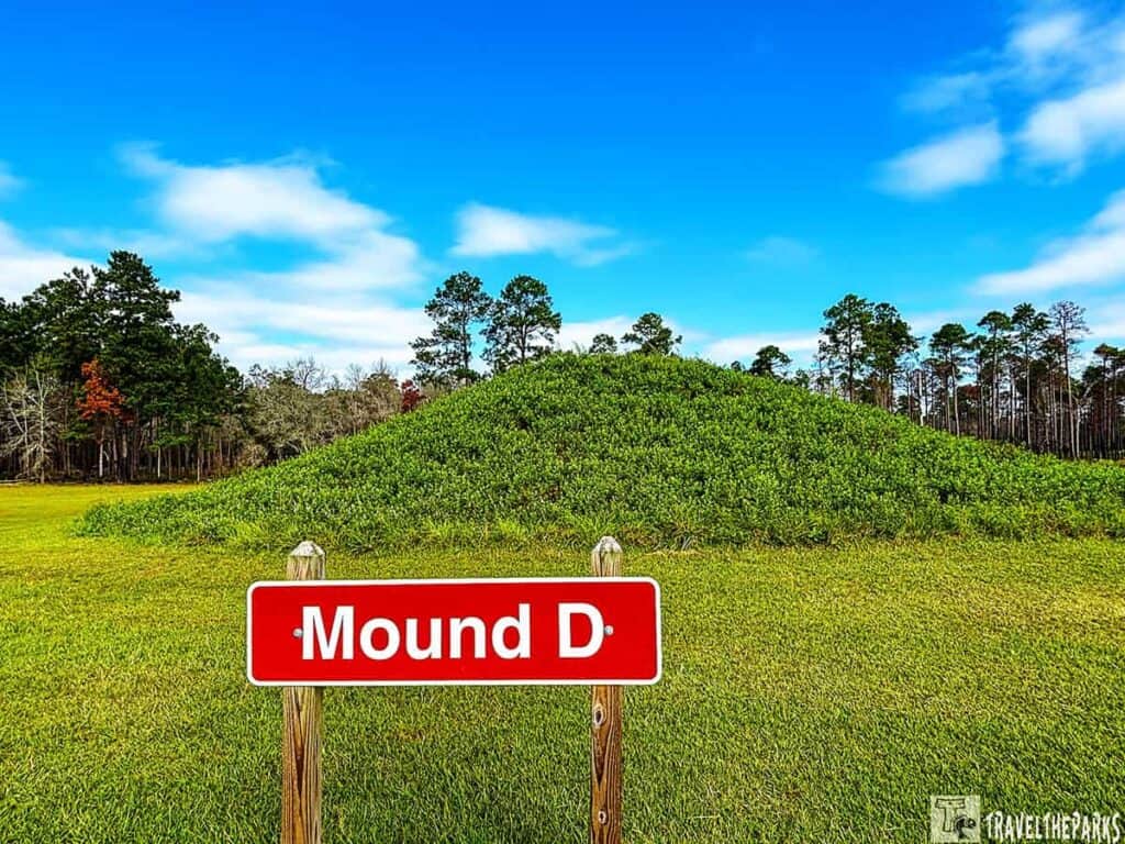A grassy mound labeled "Mound D" with a sign in a park setting, under a blue sky.

