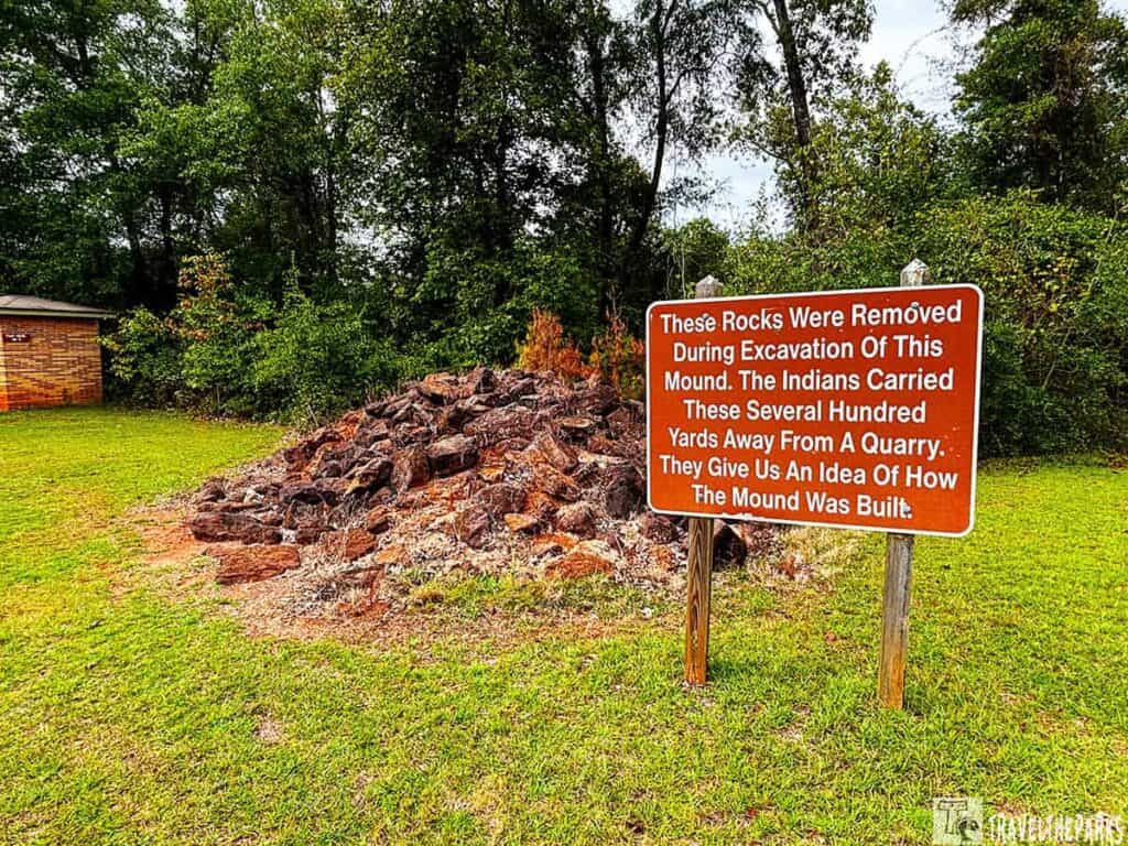 A pile of rocks with an informational sign for Mound E at Kolomoki Mounds State Park, surrounded by grass and trees.


