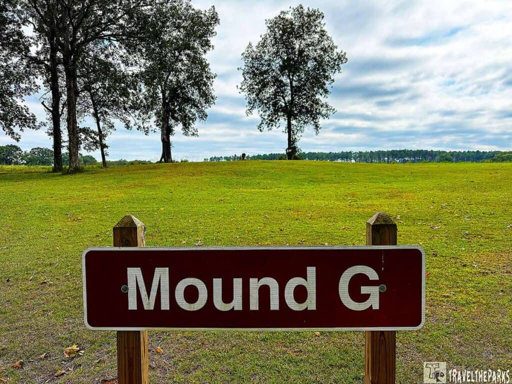Sign reading "Mound G" in front of a grassy landscape with three trees and a cloudy sky at Kolomoki Mounds State Park.

