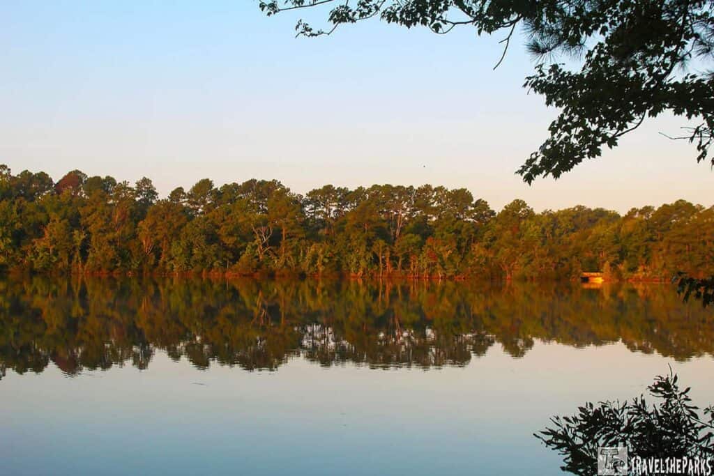 Lake reflecting trees with a small structure on the right; silhouetted branches frame the image in Virginia's historical Triangle.

