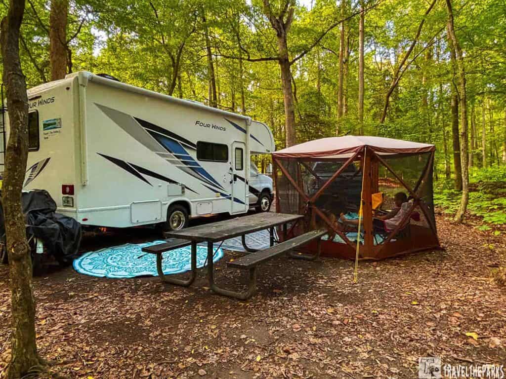Campsite with a white RV, a picnic table, and a screened tent in a forested area.