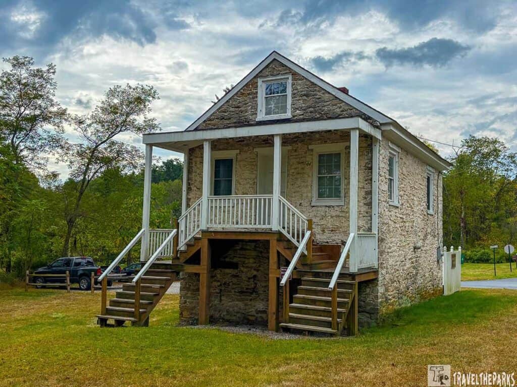 Historic stone cabin with a porch and stairs, surrounded by greenery and an overcast sky.