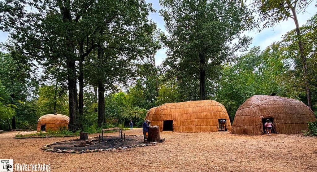 View of a Powhatan Indian village with three traditional domed structures on a dirt clearing, surrounded by trees.