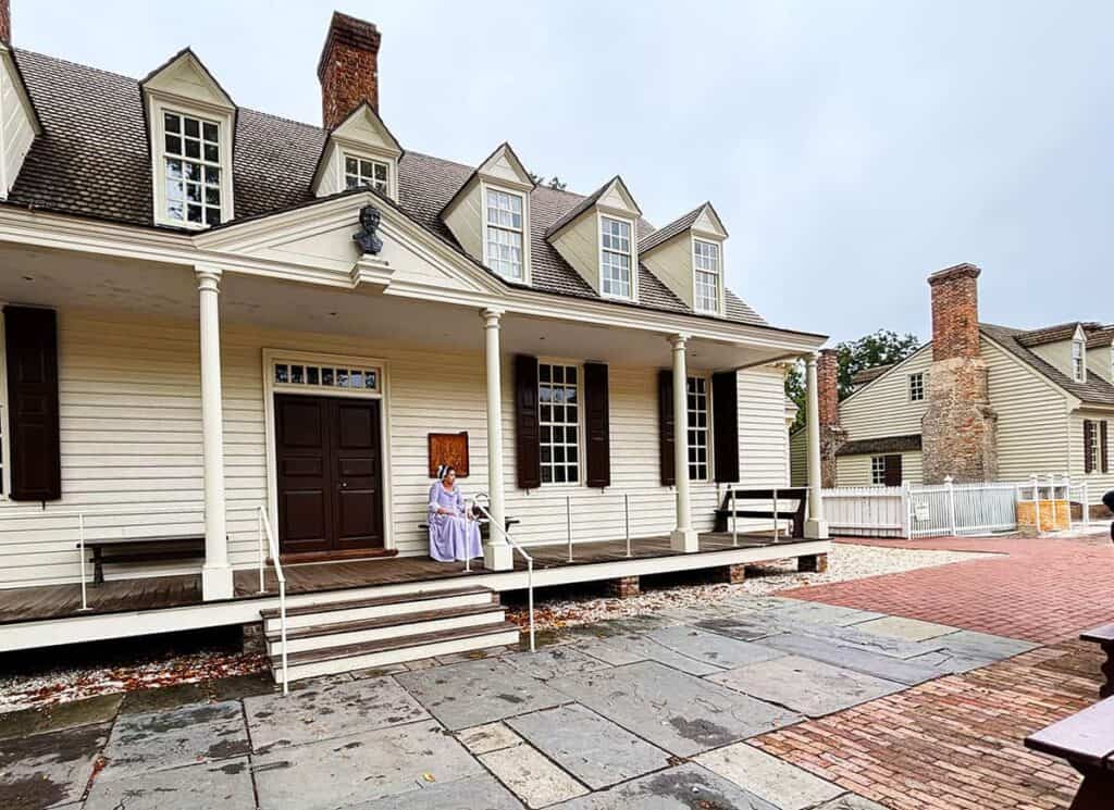 Colonial-style Raleigh Tavern with a porch, dormer windows, and a woman in period attire sitting on the steps.