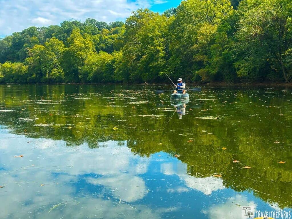 A person fishing from a kayak on a calm river in a lush, green wooded area.