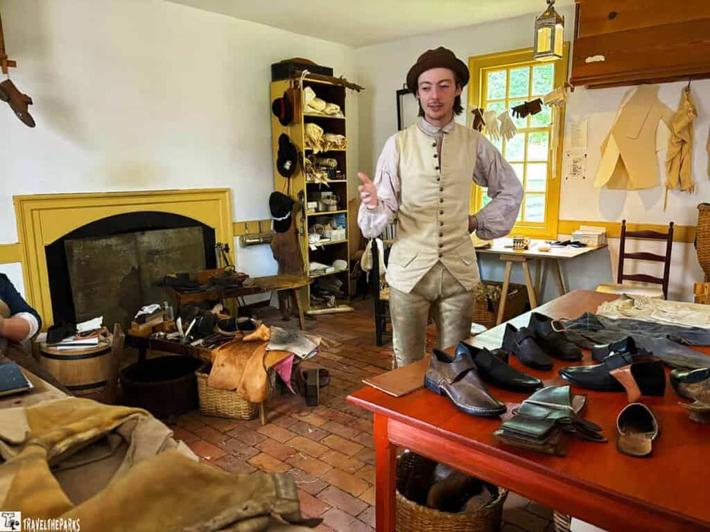 A person in period attire stands in a historical cobbler's workshop with shoes and tools on a table.