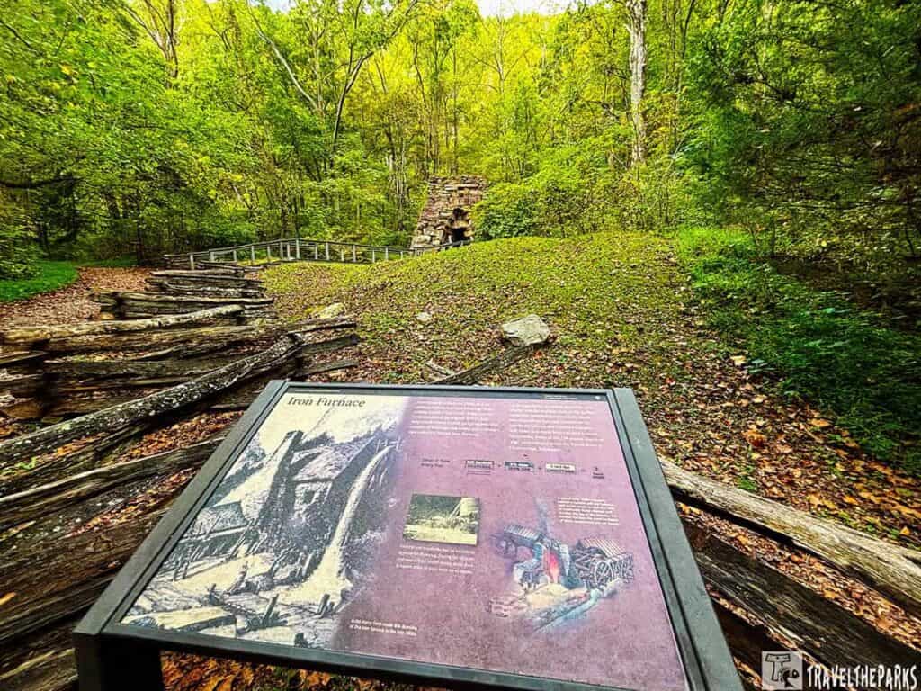 Historical iron furnace with informational sign in a lush green forest.