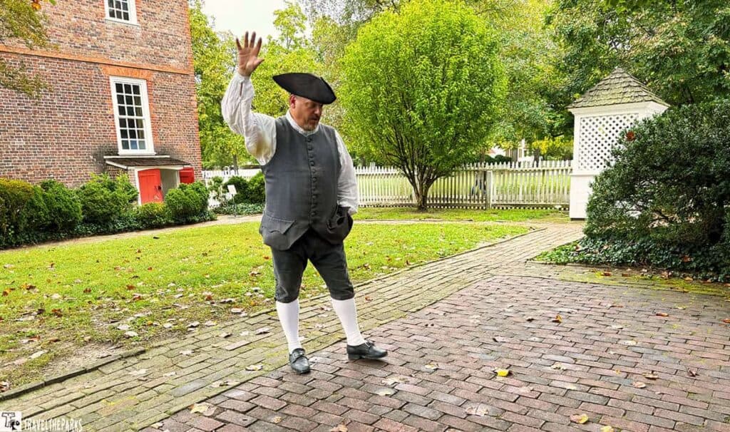 Person in historical clothing gesturing on a brick path in a garden setting near a brick building with a red door.