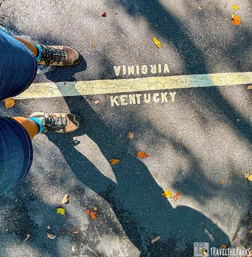Feet in hiking shoes straddle a painted line marking the Virginia-Kentucky border on an asphalt road.