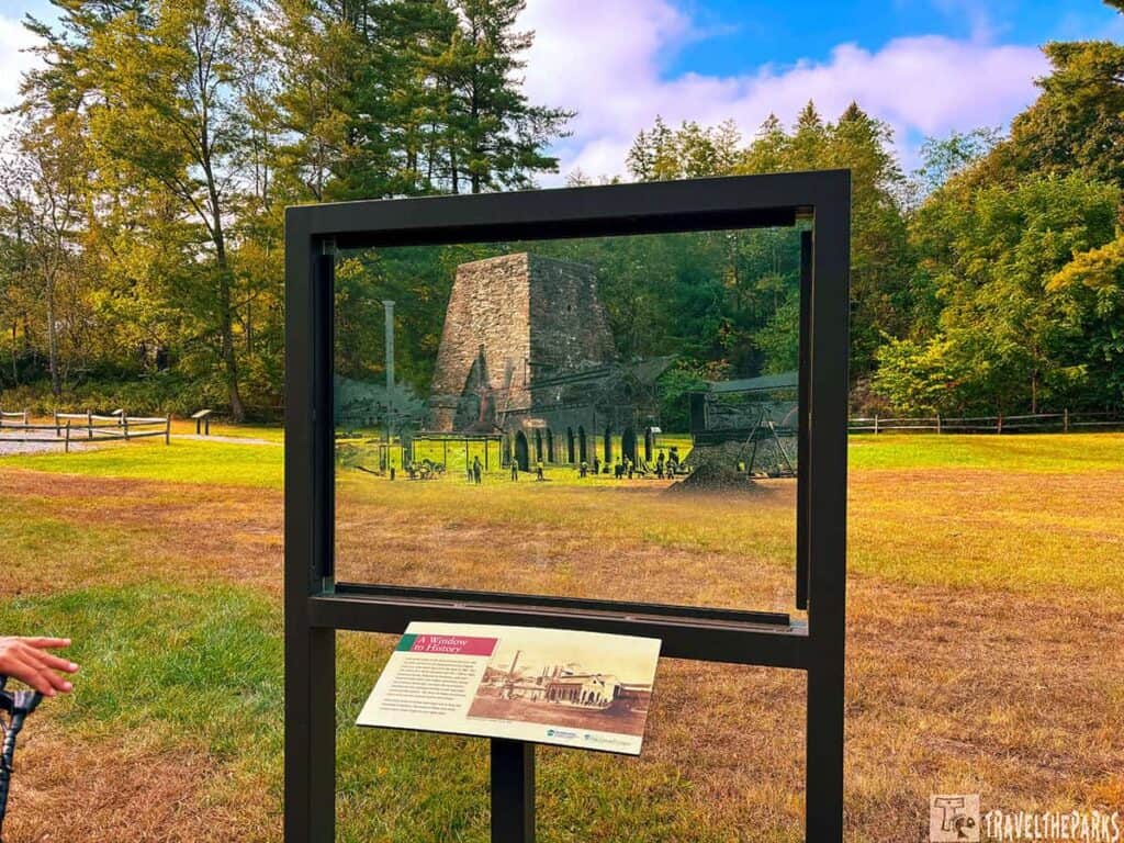 outdoor scene featuring a framed glass panel that provides a historical view overlay in a park setting. The panel displays an old image of a stone furnace, part of Pine Grove Furnace, with historical machinery and people depicted. This overlay is superimposed against the actual natural background, which consists of green grass, trees with leaves transitioning to autumn colors, and a partly cloudy blue sky. A wooden fence runs along the background, enhancing the park-like atmosphere. On the bottom right of the frame, an informational sign is attached, offering context to visitors. In the foreground, a small portion of a person's hand is visible, suggesting human interaction with the exhibit.