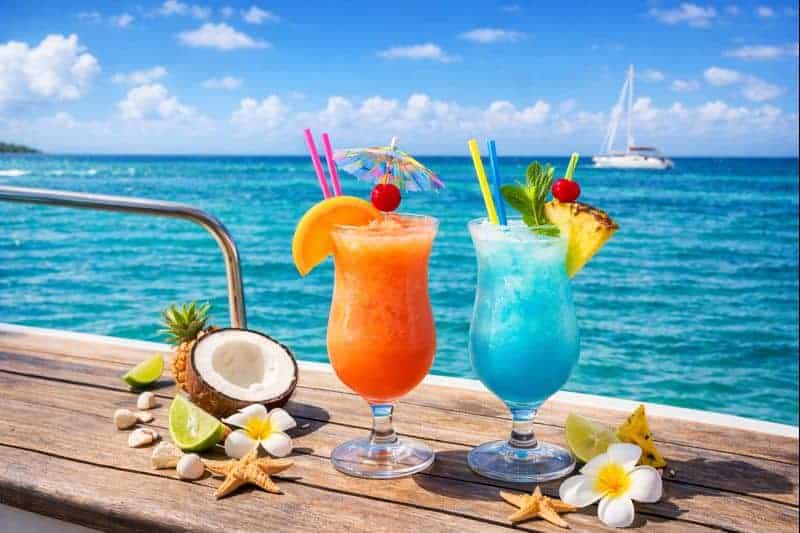 “Two colorful tropical cocktails—one orange, one blue—sit on a wooden boat deck with fruit garnishes and umbrellas, set against a bright turquoise ocean and blue sky with a sailboat in the distance.