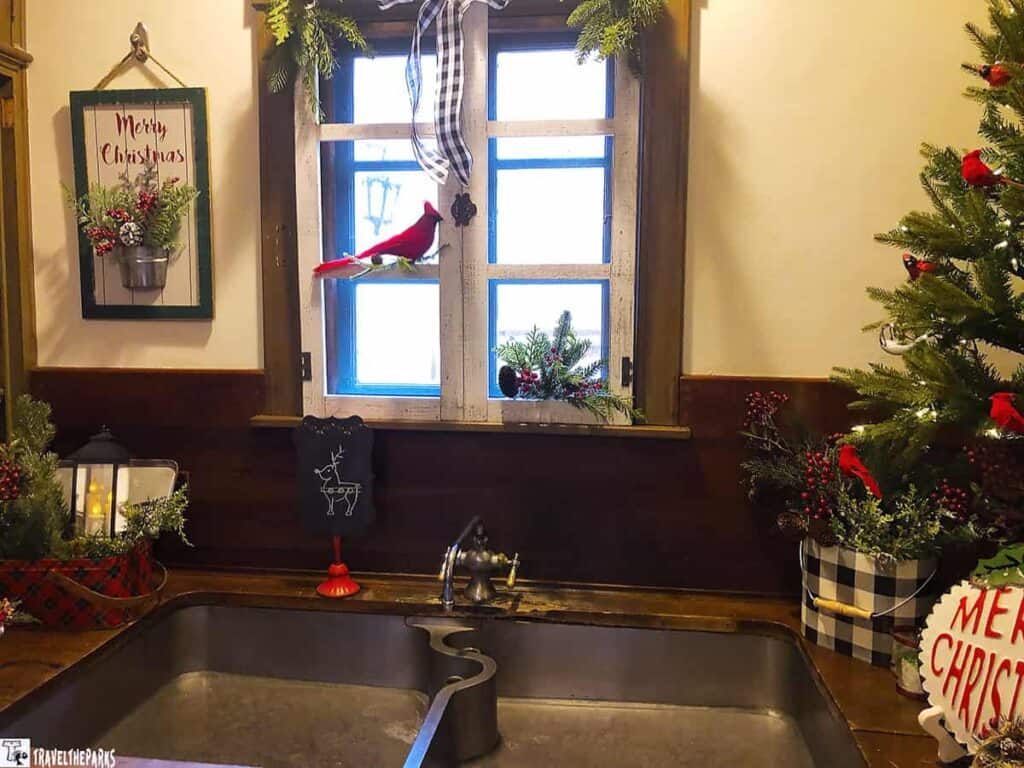 A kitchen window decorated with Christmas greenery, cardinals, and a striped ribbon. A plaid container with greenery and berries is on the right. A lantern and greenery are on the left.

