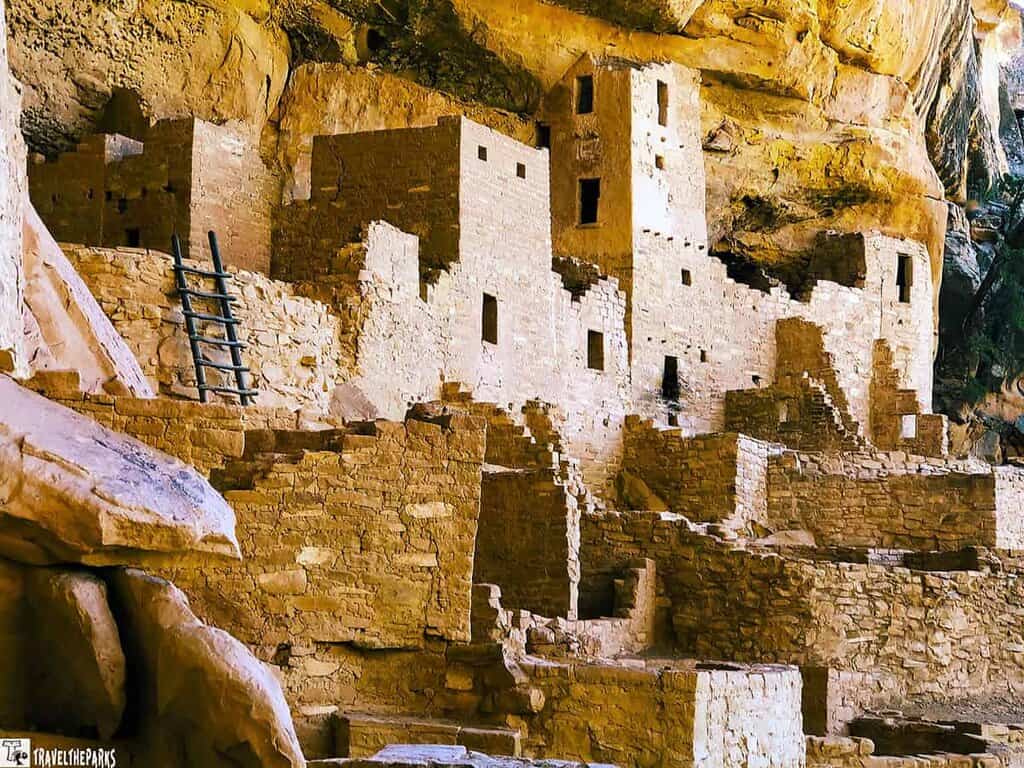 Ancient stone structures nestled in a sandstone alcove, featuring multiple stories, windows, and a wooden ladder.