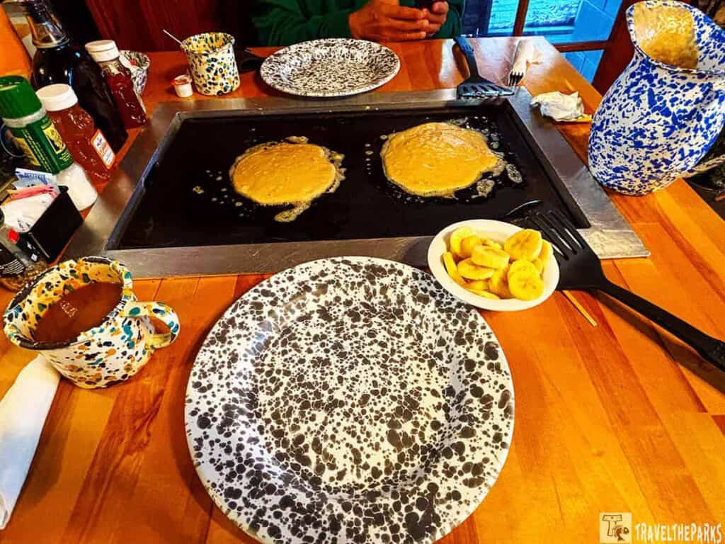A tabletop with pancakes cooking on a griddle, surrounded by speckled dishes, banana slices, condiments, and a blue speckled pitcher.