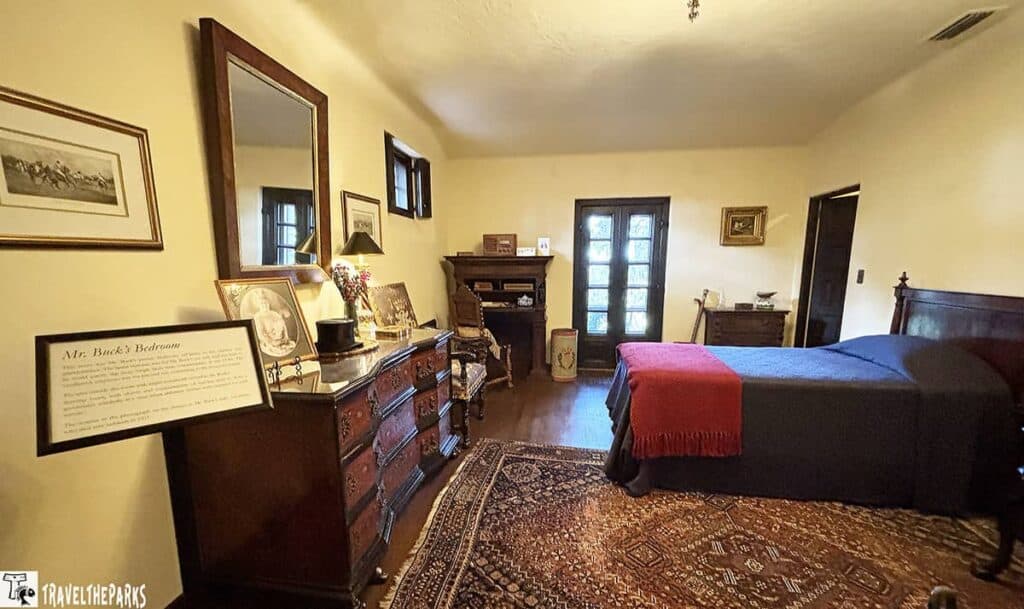 A traditionally furnished bedroom with a wooden dresser, mirror, double bed, fireplace, and glass-paneled doors.