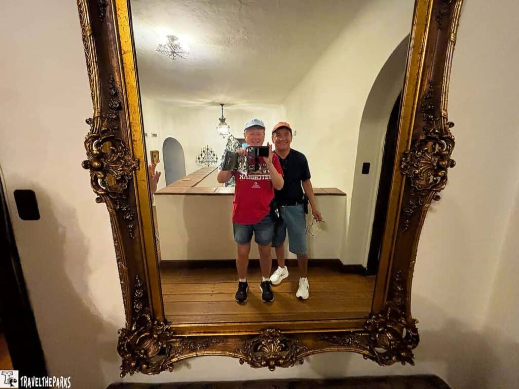 Two individuals taking a selfie in an ornate mirror with a golden frame, reflecting a room with light fixtures and an arched doorway.

