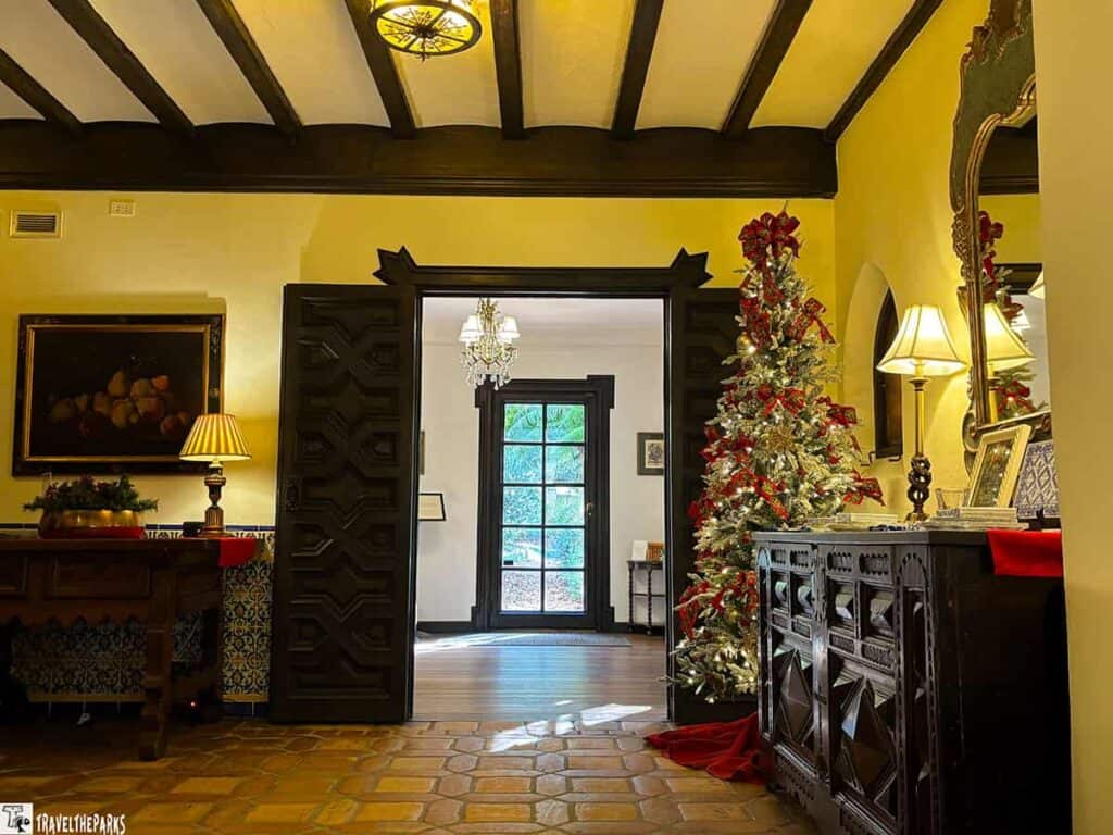 Interior of a warmly lit room with wooden beams, a Christmas tree, a wooden cabinet, and an open doorway leading to another room.


