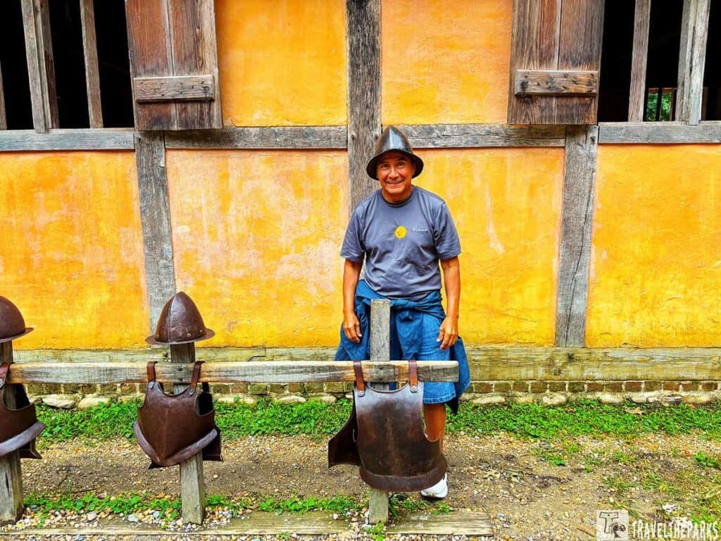 A person stands in front of a historic building with yellow-orange walls and wooden beams at Jamestown Settlement's James Fort, with armor displayed on a wooden fence.


