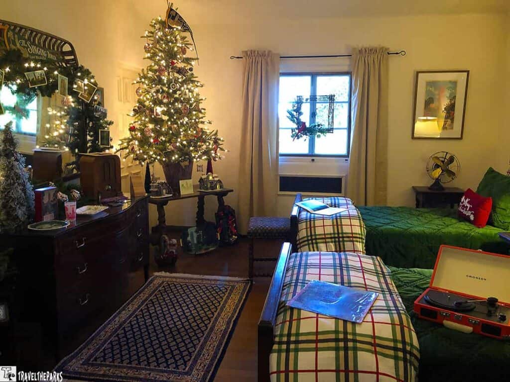 A cozy room with a lit Christmas tree, vintage dresser, and plaid-adorned twin beds decorated for the holidays.

