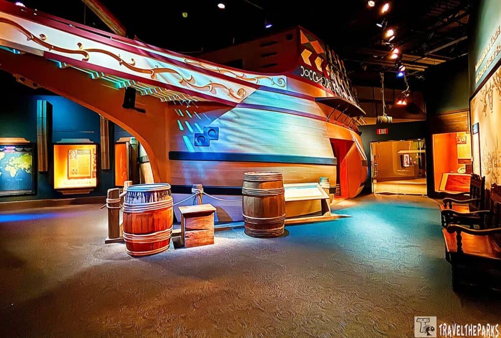 Interior view of the Jamestown Settlement Museum featuring a life-sized ship hull replica with barrels and a chest displayed in the foreground.
