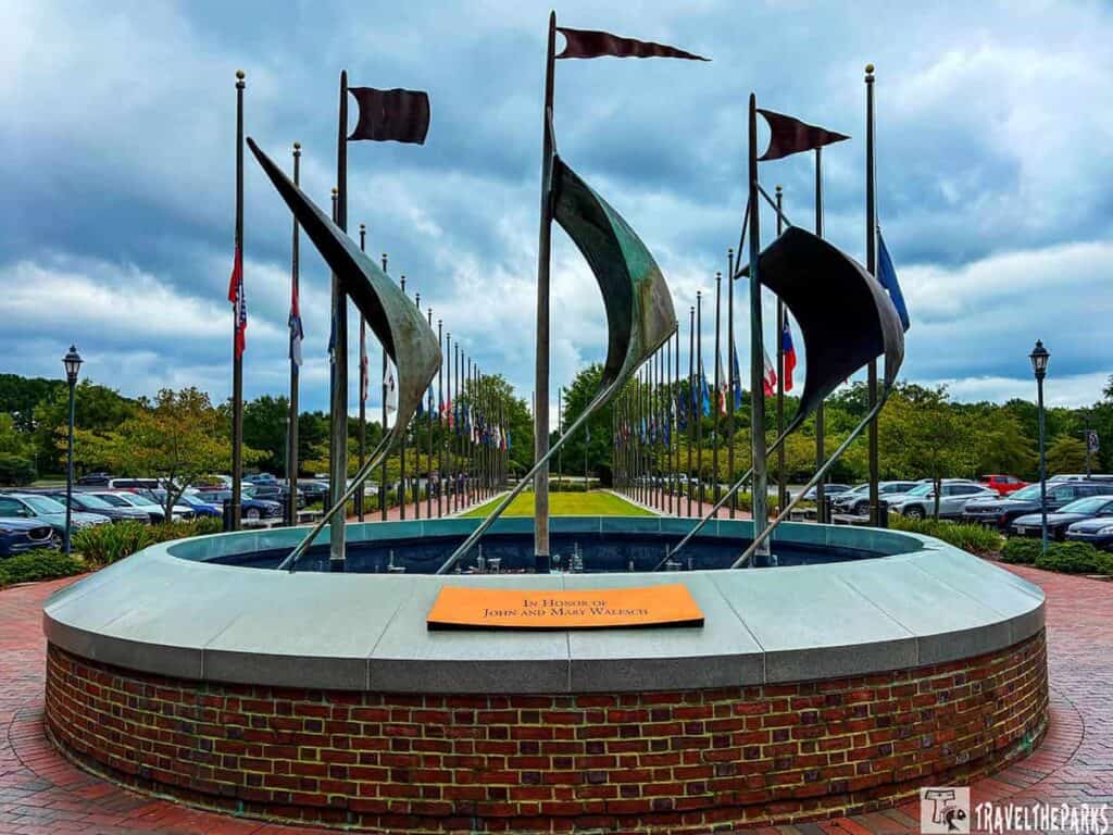 Jamestown Settlement outdoor display with sail-like sculptures and flags lining a walkway under a cloudy sky.

