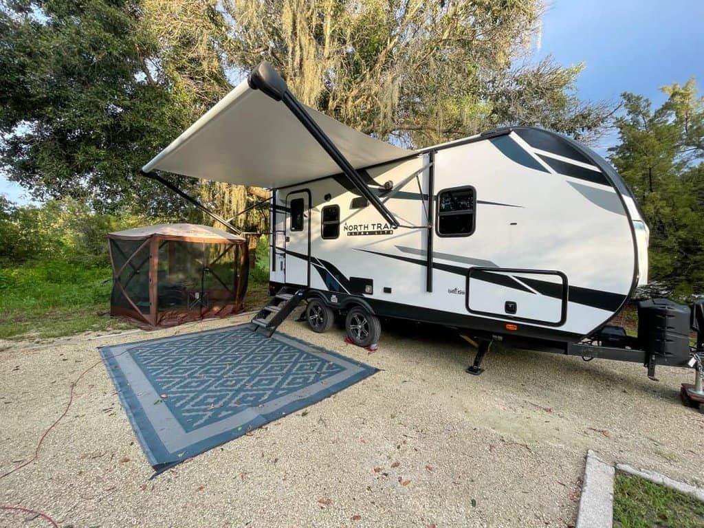 Travel trailer at a campsite with an extended awning, outdoor rug, and screened tent.