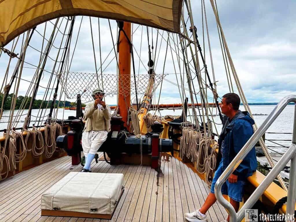 Two people on the deck of a historic sailing ship with ropes and rigging, one in period costume and the other in modern attire.

