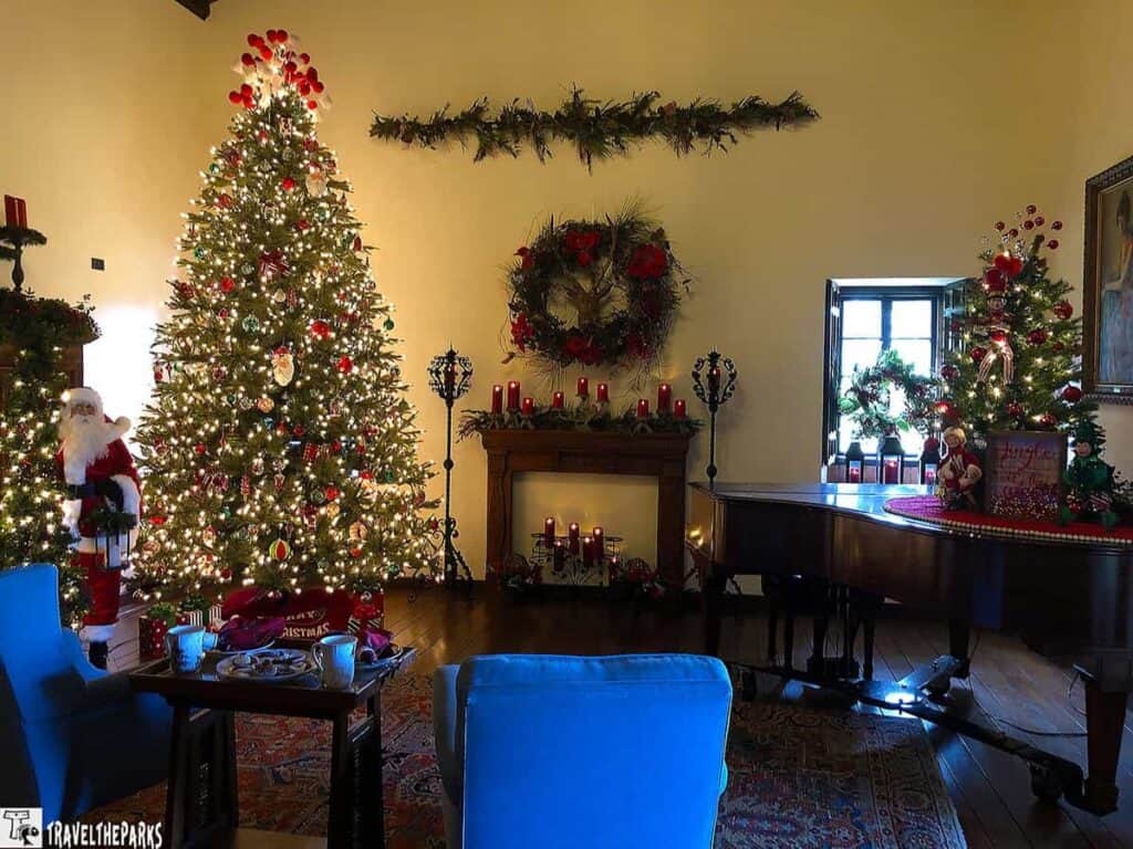 A living room decorated for Christmas with a large tree, a fireplace, and a grand piano, adorned with festive ornaments and lights.


