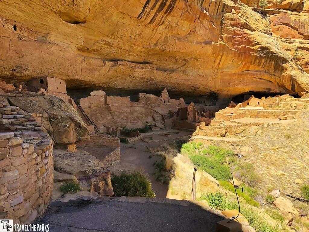 Ruins of Long House at Mesa Verde under an overhanging orange and brown cliff.

