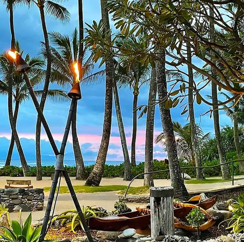 Mammas Fish House-A Hawaiian landscape with lit tiki torches, palm trees, and a colorful sky at sunset.