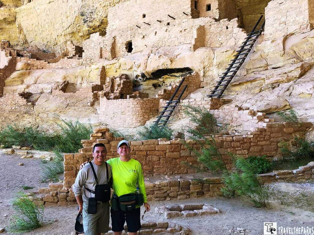 Two people stand in front of an ancient Longhouse cliff dwelling with stone structures and wooden ladders.