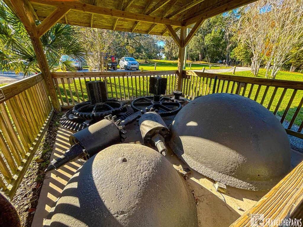 Industrial machinery parts displayed under a wooden pavilion in a park setting.

