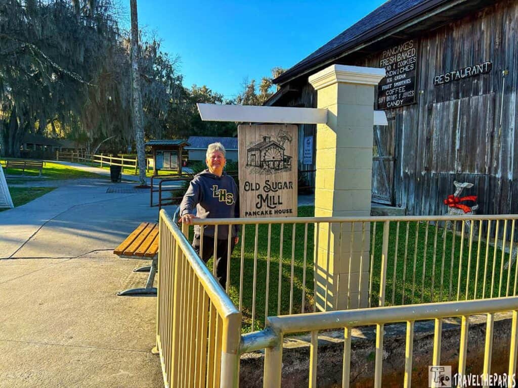 Person standing next to the "Old Sugar Mill Pancake House" sign at De Leon Springs, with a rustic building and trees in the background.

