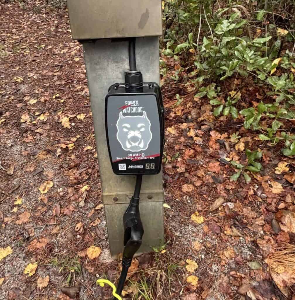 Black "Power Watchdog" surge protector connected to a tall metal post amidst fallen leaves and foliage.