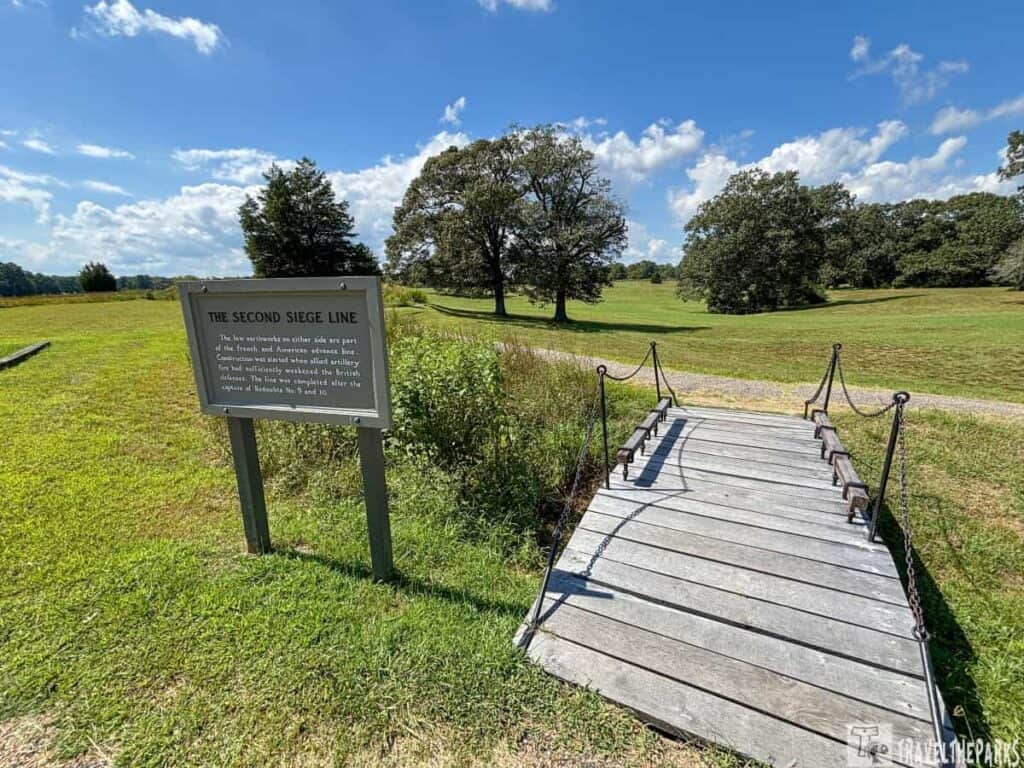 View of Yorktown National Battlefield with a sign about the "Second Siege Line" and a wooden bridge leading to a gravel path.