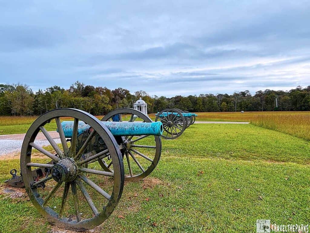 Row of historic cannons on a grassy field at the Chickamauga Battlefield with a white monument in the background.

