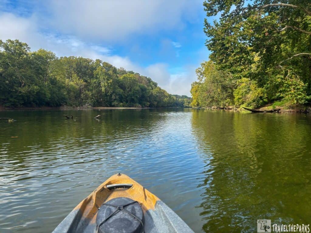 Kayak view of a calm Shenandoah River State park surrounded by green trees under a partly cloudy sky.